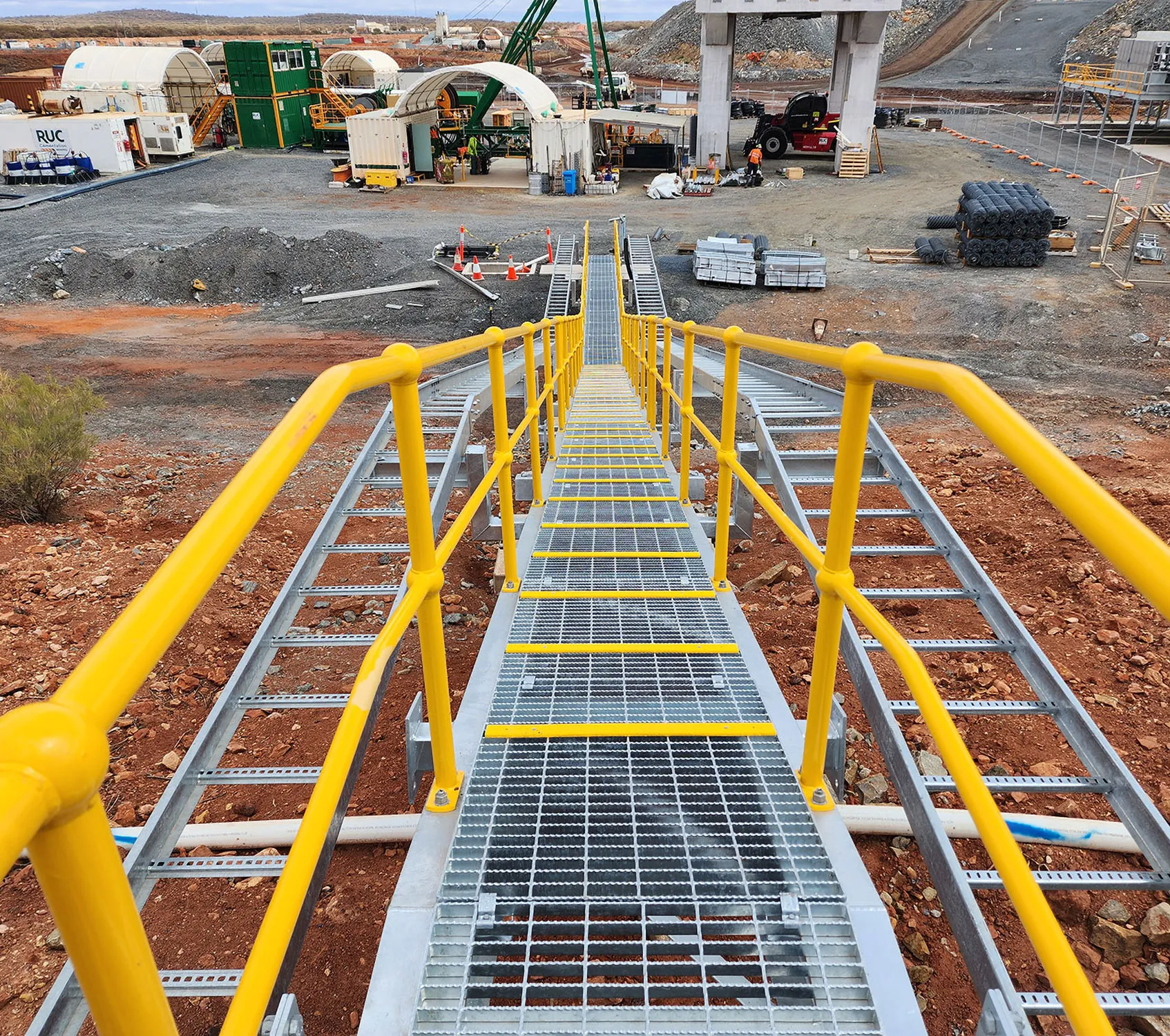 Elevated steel walkway with yellow handrails leading down to a mine site compound of site offices and equipment